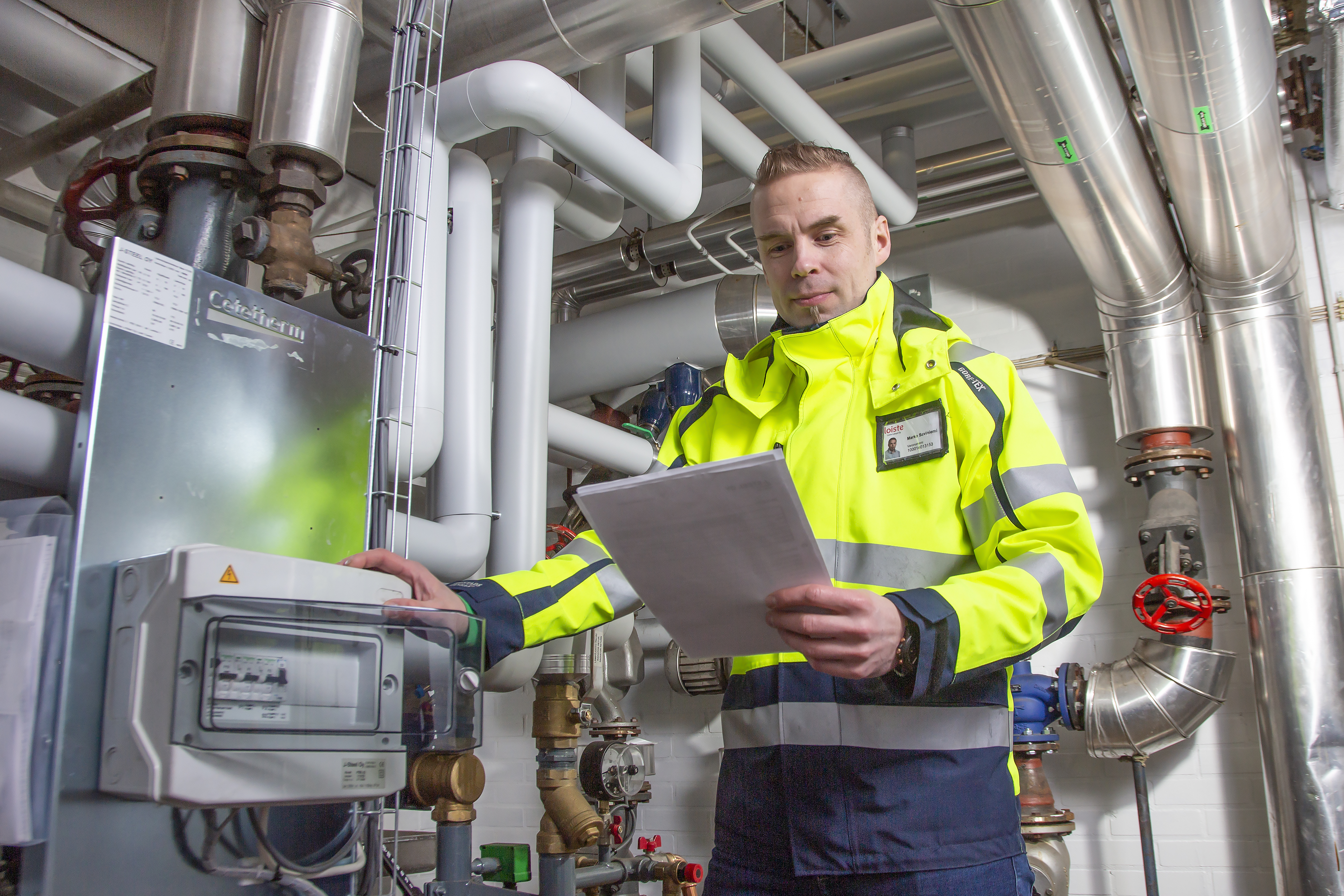 Kuvassa Marko Saviniemi lämmönjakohuoneessa paperit kädessään ja huomiovärivaatteet päällä.